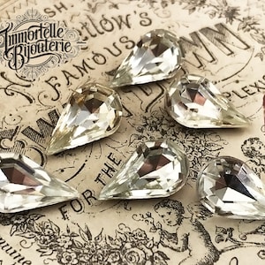 May include: Six clear, teardrop-shaped rhinestones with multiple facets are arranged on a vintage paper background. The rhinestones are highly reflective, catching the light. The image includes the text "Immortelle Bijouterie".