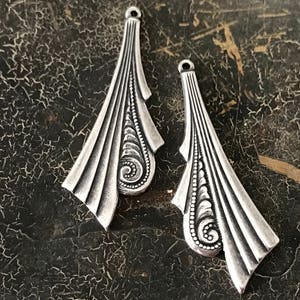 May include: Two silver metal pendant charms with an ornate design. The charms are shaped like a teardrop with a pointed tip and a rounded bottom. The design features a series of curved lines and swirls.