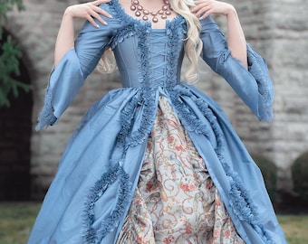 Könnte beinhalten: Ein puderblaues historisches Kleid mit Rüschenmieder und langen Ärmeln. Das Kleid hat einen floral gemusterten Unterrock und ein geschnürtes Korsett. Das Kleid erinnert an die Mode des 18. Jahrhunderts.