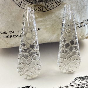 May include: Two clear, teardrop-shaped glass pendants with a textured, honeycomb-like pattern. The pendants are displayed on a white surface with a vintage paper background. The pendants are likely for jewelry making.