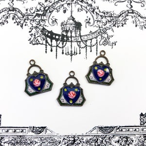 May include: Three antique-style metal charms in the shape of a heart-shaped purse with a blue enamel background and a pink rose design.
