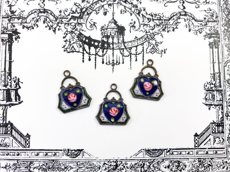 May include: Three antique-style metal charms in the shape of a heart-shaped purse with a blue enamel background and a pink rose design.