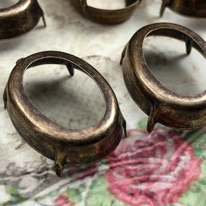 May include: Oval-shaped metal settings with a brass finish and three prongs on the bottom. The settings are designed to hold a cabochon or other flat-backed stone.