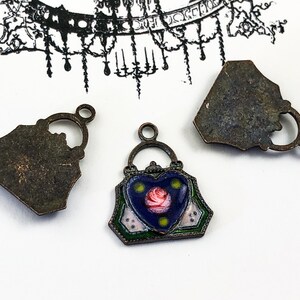 May include: Three antique bronze-colored metal charms in the shape of a handbag. The charm in the center has a blue enamel background with a pink rose and green and white accents. The other two charms are the back of the handbag charm.