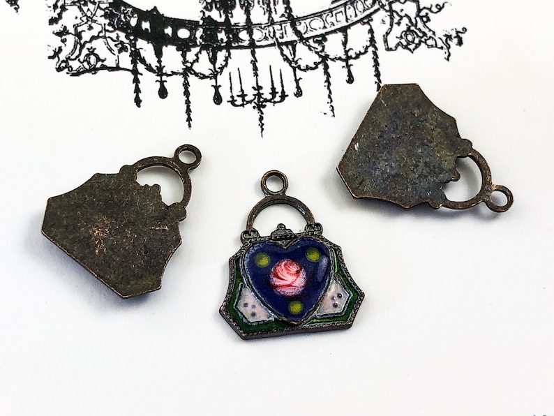 May include: Three antique bronze-colored metal charms in the shape of a handbag. The charm in the center has a blue enamel background with a pink rose and green and white accents. The other two charms are the back of the handbag charm.