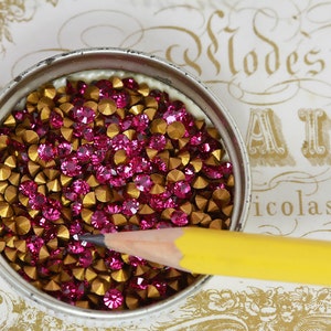 May include: A close-up of a small tin container filled with pink and gold rhinestones. The container is sitting on a white surface with a decorative pattern.