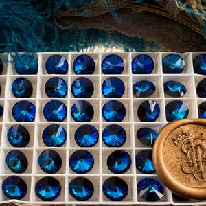 K&ouml;nnte beinhalten: Eine Ansammlung von facettierten, runden, dunkelblauen Kristallen mit schillerndem Finish, angeordnet in einem wei&szlig;en Raster. Ein goldfarbenes Wachssiegel mit einem verzierten Emblem befindet sich unten rechts. Geeignet f&uuml;r Schmuckherstellung.
