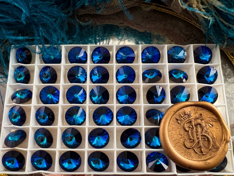K&ouml;nnte beinhalten: Eine Ansammlung von facettierten, runden, dunkelblauen Kristallen mit schillerndem Finish, angeordnet in einem wei&szlig;en Raster. Ein goldfarbenes Wachssiegel mit einem verzierten Emblem befindet sich unten rechts. Geeignet f&uuml;r Schmuckherstellung.