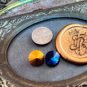 K&ouml;nnte beinhalten: Anordnung dekorativer Elemente, darunter ein goldfarbenes Wachssiegel mit Kronen- und Initialendesign, zwei facettierte Edelsteine in Gold und Blau sowie eine US-Dime-M&uuml;nze. Die Elemente sind in einem verzierten, ovalen Rahmen angeordnet.