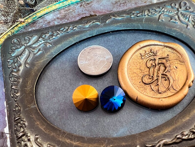 K&ouml;nnte beinhalten: Anordnung dekorativer Elemente, darunter ein goldfarbenes Wachssiegel mit Kronen- und Initialendesign, zwei facettierte Edelsteine in Gold und Blau sowie eine US-Dime-M&uuml;nze. Die Elemente sind in einem verzierten, ovalen Rahmen angeordnet.