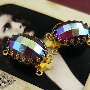 May include: Two gold-tone metal clasps with oval-shaped iridescent purple and blue glass stones. The stones have a faceted design.