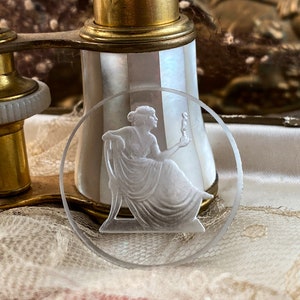 May include: A clear glass round coaster with a raised, frosted design of a woman holding a torch. The coaster is sitting on a white lace surface.