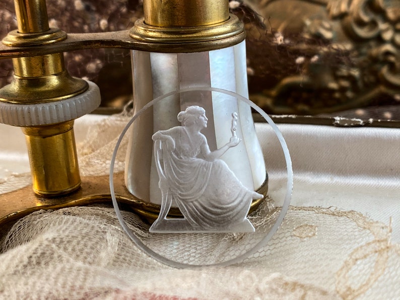May include: A clear glass round coaster with a raised, frosted design of a woman holding a torch. The coaster is sitting on a white lace surface.