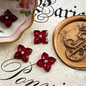 May include: Four red, four-pointed star-shaped embellishments with a clear rhinestone centre. The embellishments are scattered on a white surface with a gold wax seal in the background.