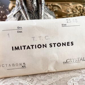 Puede incluir: Una bolsa de papel blanca con el texto "IMITATION STONES" impreso en negro. La bolsa tambi&eacute;n tiene el texto "OCTAGONS Art." y "Col. CRYSTAL" impreso en ella. La bolsa tiene un n&uacute;mero escrito a mano "1" y el texto "Grs. Doz. T.T.C. Size 25/18" impreso en ella.