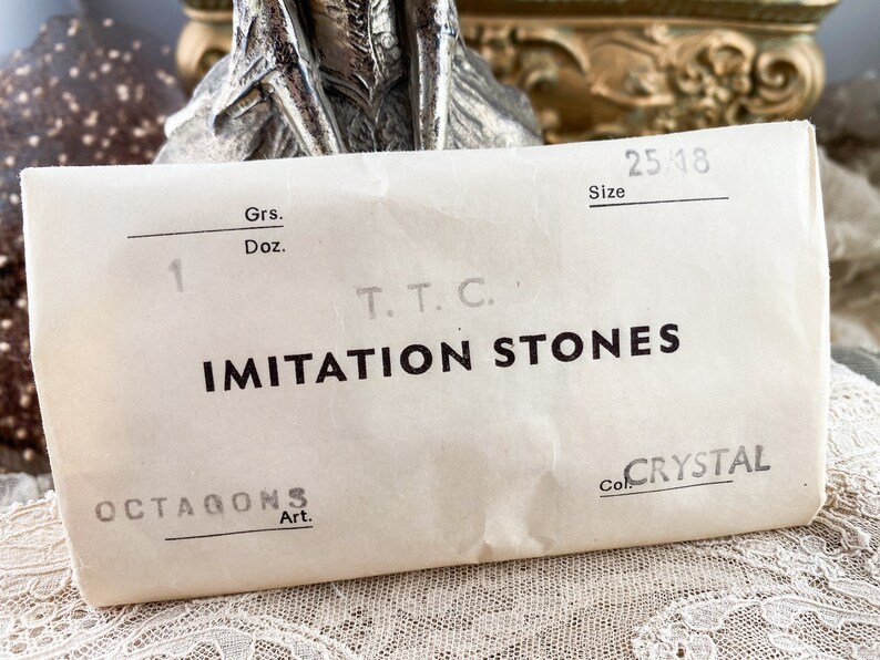 Puede incluir: Una bolsa de papel blanca con el texto "IMITATION STONES" impreso en negro. La bolsa tambi&eacute;n tiene el texto "OCTAGONS Art." y "Col. CRYSTAL" impreso en ella. La bolsa tiene un n&uacute;mero escrito a mano "1" y el texto "Grs. Doz. T.T.C. Size 25/18" impreso en ella.