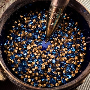 May include: A close-up of a small, round, brass container with a blue interior. The container is filled with gold and blue rhinestones. A gold-coloured pen nib is partially inserted into the container.