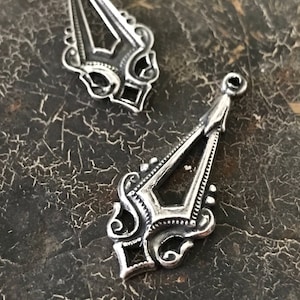 May include: Two silver-toned metal pendant charms with intricate, ornate designs. The charms are shaped like a pointed teardrop with a decorative openwork pattern.