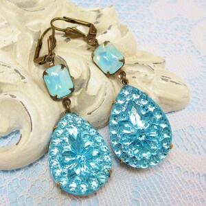 May include: A pair of dangle earrings featuring a teardrop-shaped blue glass stone and a smaller square-shaped opal stone. The earrings have a gold-tone finish.