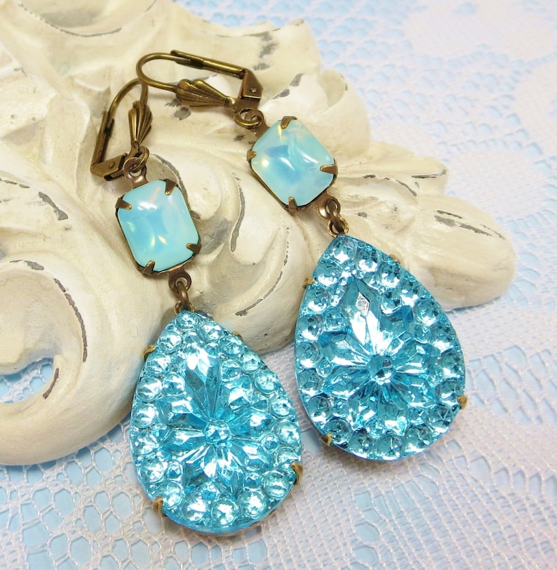 May include: A pair of dangle earrings featuring a teardrop-shaped blue glass stone and a smaller square-shaped opal stone. The earrings have a gold-tone finish.