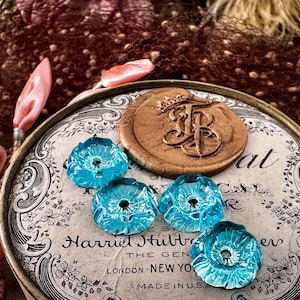 May include: A vintage tin box with a floral design and the text "Harriet Hubbard Ayer The Genuine London New York Made in U.S.A."  The box has a wax seal with a crown and the letter "F" on top.  Four turquoise blue glass flower-shaped beads are on top of the box.