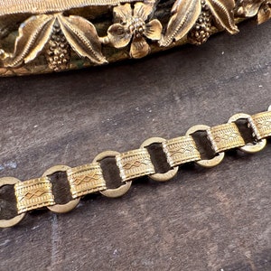 K&ouml;nnte beinhalten: Ein goldfarbenes Kettenarmband mit einem dekorativen, gemusterten Design. Die Kette besteht aus miteinander verbundenen Kreisen mit einem strukturierten, gemusterten Band.