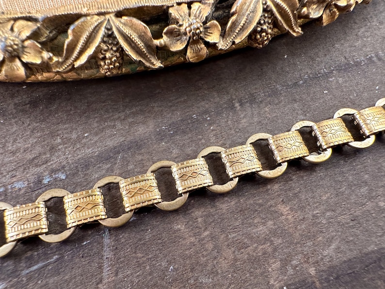 K&ouml;nnte beinhalten: Ein goldfarbenes Kettenarmband mit einem dekorativen, gemusterten Design. Die Kette besteht aus miteinander verbundenen Kreisen mit einem strukturierten, gemusterten Band.