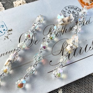 May include: Two long, dangling crystal necklaces with iridescent beads and silver chains. The necklaces are on a white surface with a vintage postcard.