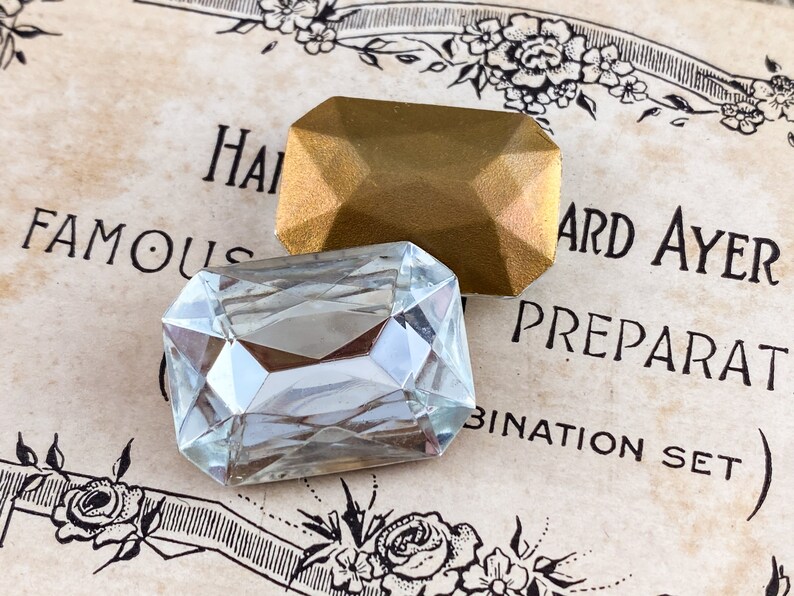 Puede incluir: Dos gemas de vidrio facetadas, una transparente y otra dorada, sobre un fondo de papel vintage con el texto "Famous Hard Ayer Preparat Combination Set".