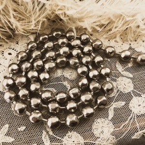 May include: A silver beaded garland with a metallic finish. The beads are round and have a slightly textured surface. The garland is coiled and lying on a white lace fabric.
