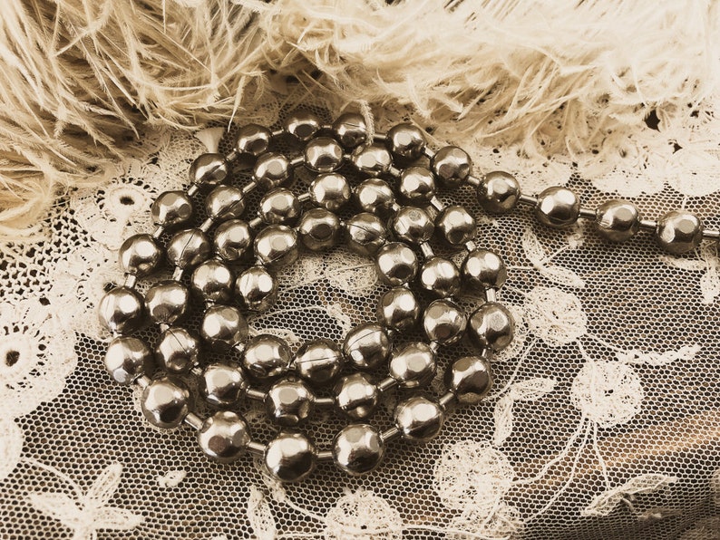 May include: A silver beaded garland with a metallic finish. The beads are round and have a slightly textured surface. The garland is coiled and lying on a white lace fabric.