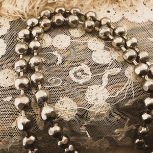 May include: A silver beaded necklace draped over a delicate white lace fabric. The beads are round and have a slightly textured surface.