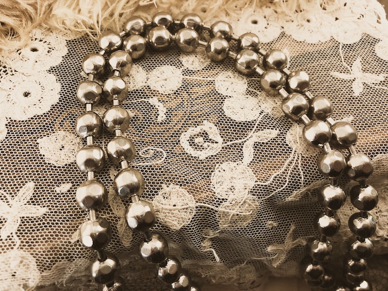 May include: A silver beaded necklace draped over a delicate white lace fabric. The beads are round and have a slightly textured surface.