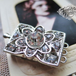 May include: Silver-toned jewelry clasp with a square, floral-inspired design. The clasp is adorned with multiple clear, sparkling crystals. The design includes a large central crystal and smaller crystals around the edges.