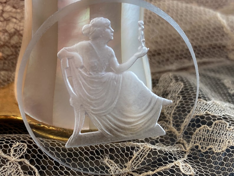 May include: A white glass cameo featuring a woman sitting on a chair, holding a small figure in her hand. The cameo is set in a clear glass circle.
