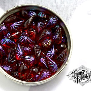 May include: A bowl filled with iridescent red glass beads with a starburst pattern. The beads have a purple and blue iridescent sheen.