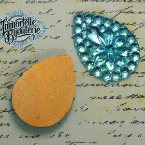 May include: A turquoise blue glass teardrop-shaped embellishment with a raised, textured design. The back of the embellishment is a flat, gold-coloured surface.