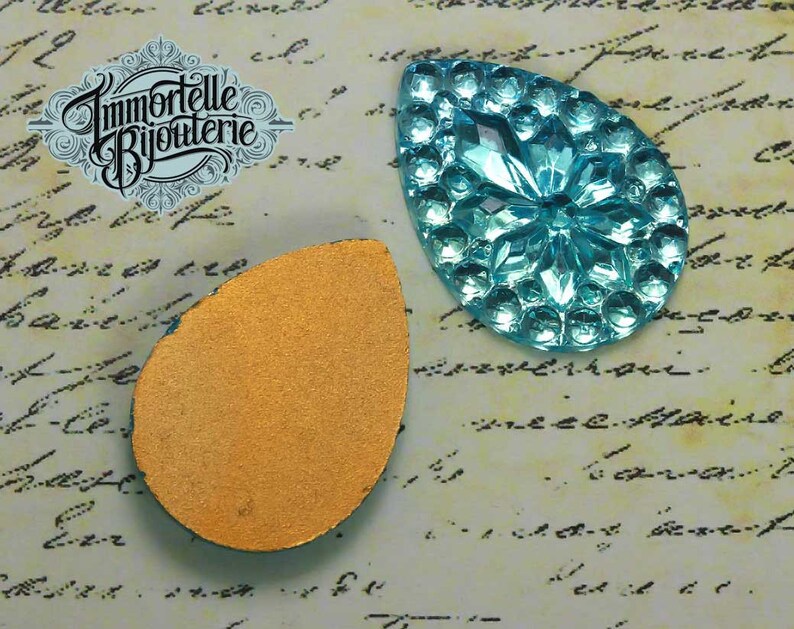 May include: A turquoise blue glass teardrop-shaped embellishment with a raised, textured design. The back of the embellishment is a flat, gold-coloured surface.