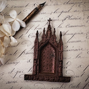 May include: A detailed, antique-style, brown miniature church or shrine with a cross on top. The decorative piece sits on a vintage paper background with handwritten script, alongside a dried flower arrangement and a pen.