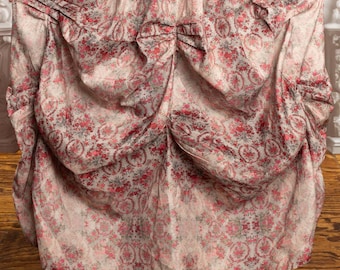 May include: A full-length skirt with a floral pattern in shades of pink, red, and gray on a cream background. The skirt is gathered at the waist and has a draped, layered design. The fabric appears to be lightweight.