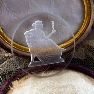 May include: A white cameo with a raised design of a woman holding a small object. The cameo is set in a gold-colored frame.