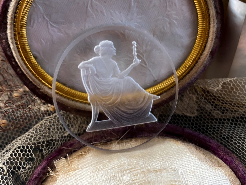 May include: A white cameo with a raised design of a woman holding a small object. The cameo is set in a gold-colored frame.
