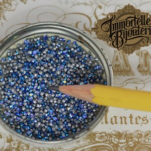 May include: A close-up of a small, round container filled with blue and silver rhinestones. A yellow pencil is lying on top of the rhinestones.