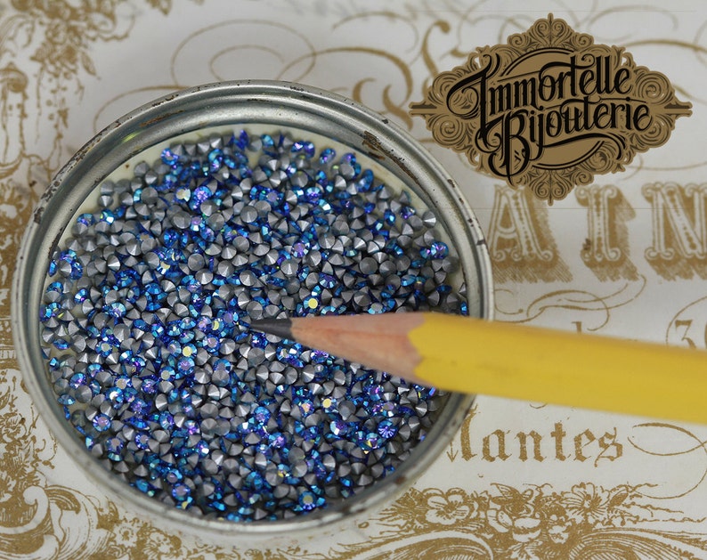 May include: A close-up of a small, round container filled with blue and silver rhinestones. A yellow pencil is lying on top of the rhinestones.