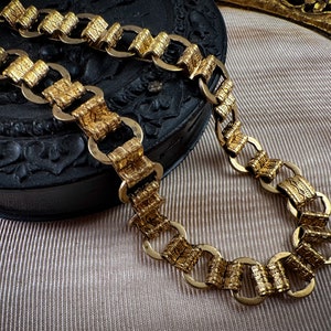 K&ouml;nnte beinhalten: Eine goldene Kettenkette mit einem strukturierten, verzierten Design. Die Kette besteht aus ineinandergreifenden ovalen Gliedern.