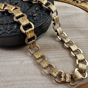 K&ouml;nnte beinhalten: Eine goldene Kettenkette mit einem detaillierten, verzierten Design. Die Kette besteht aus miteinander verbundenen Kreisen mit komplizierten Mustern.