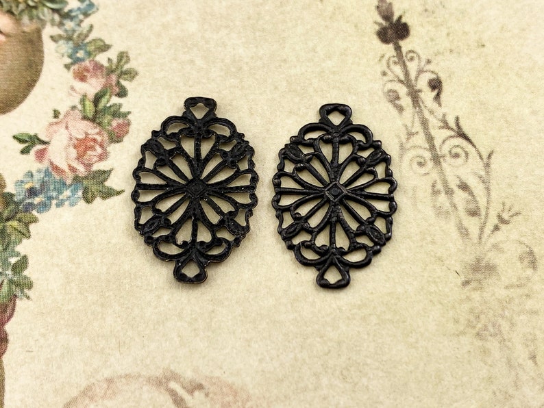 Puede incluir: Dos abalorios de metal filigranado negro en forma de flor con un centro circular. Los abalorios son de forma ovalada y tienen un dise&ntilde;o delicado e intrincado.