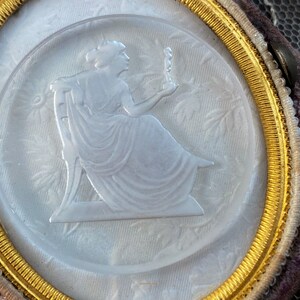 May include: A white cameo with a gold rim, featuring a woman in a flowing gown holding a small figure. The cameo is set in a velvet-covered frame.