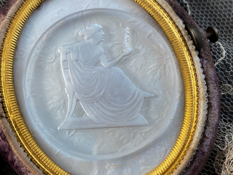May include: A white cameo with a gold rim, featuring a woman in a flowing gown holding a small figure. The cameo is set in a velvet-covered frame.
