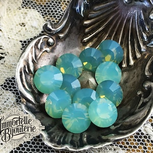 May include: A collection of pale green iridescent glass rhinestones in a silver dish. The rhinestones have a faceted design and are arranged in a cluster.